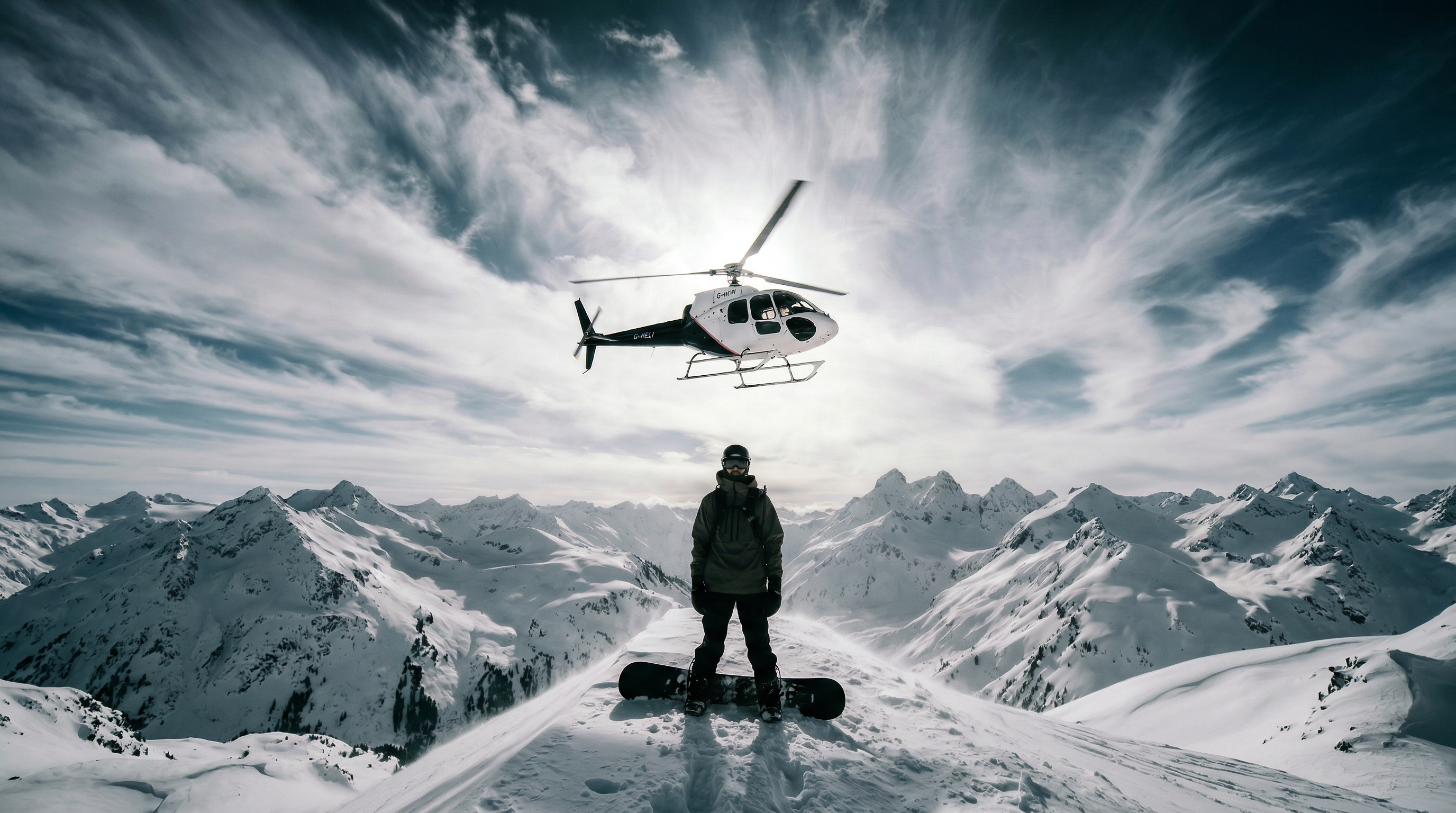 Heliboarder standing on a mountain peak with helicopter overhead and panoramic snow-covered mountains